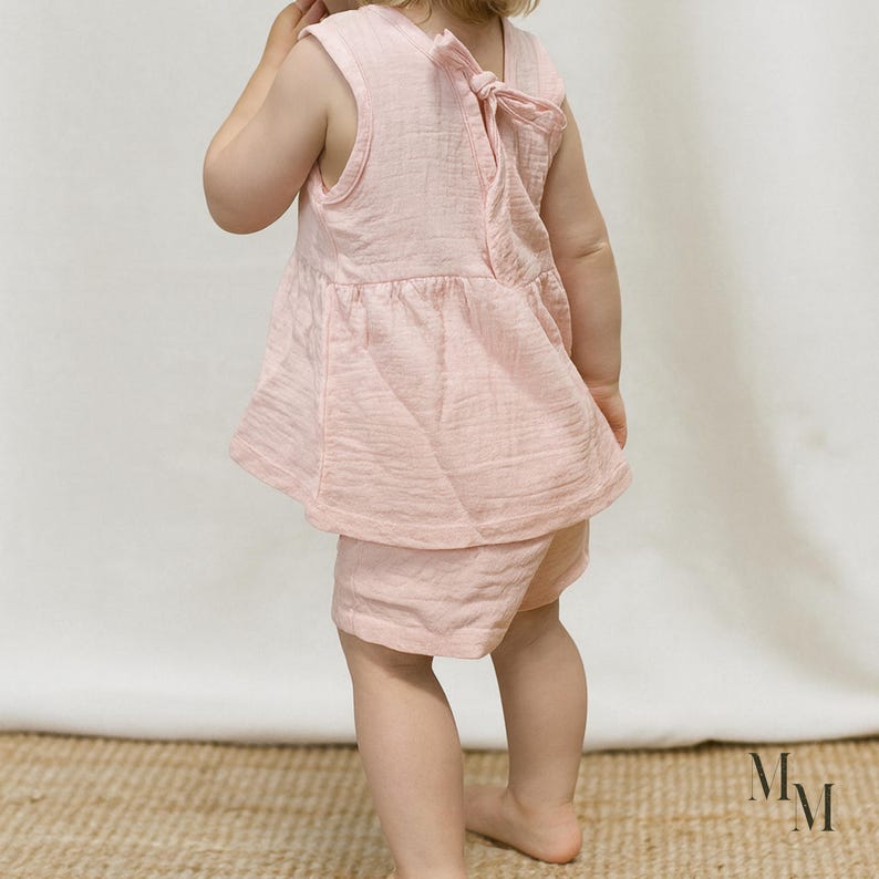 May include: A light pink baby outfit, featuring a sleeveless top with a bow detail on the back and matching shorts. The outfit is made of a textured fabric. The baby is standing on a woven mat.