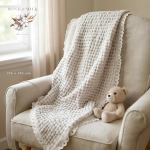May include: A light gray, textured blanket with a delicate lace trim is draped over a cream-colored armchair. A small teddy bear sits on the chair. The blanket measures 150 x 150 cm.