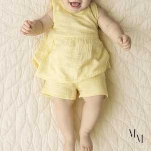 May include: A baby wearing a yellow sleeveless top and matching shorts. The outfit is made of a textured fabric. The baby is lying on a cream-colored quilted blanket. The blanket has a leaf-like pattern.