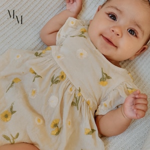 Könnte beinhalten: Ein Baby in einem beigefarbenen Kleid mit gelben und weißen Blumenstickereien. Das Kleid hat kurze Ärmel und eine geraffte Taille. Das Baby liegt auf einer hellblauen Decke und lächelt. Das Kleid ist aus einem weichen, leichten Stoff.