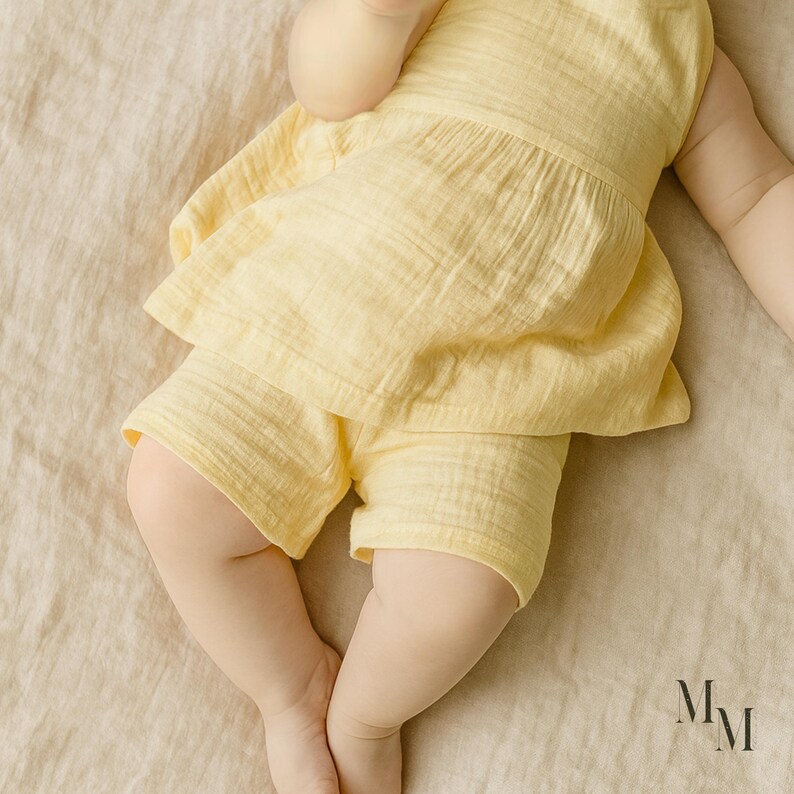May include: A baby wearing a yellow two-piece outfit, consisting of a ruffled top and shorts. The outfit is made of a textured fabric. The baby is lying on a beige surface. The letters "Mm" are visible in the bottom right corner.