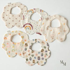 Peut inclure: Cinq bavoirs pour bébés avec un bord festonné et une ouverture de cou circulaire. Les bavoirs présentent divers motifs, notamment des cœurs, des arcs-en-ciel, des koalas et des motifs floraux. Les bavoirs sont en tissu doux et texturé aux tons neutres.