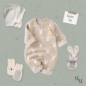 Puede incluir: Un conjunto de Pascua para bebés, con un pelele beige con estampados de conejos blancos, un gorro a juego, calcetines y un sonajero con forma de conejo. El pelele tiene mangas largas y cierres de botón. El texto "Easter Set" está en una nota pequeña.