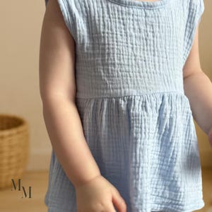 May include: A light blue, sleeveless baby dress with a textured, crinkled fabric. The dress has a round neckline and a gathered waist, creating a flowing silhouette. The dress is made of a soft, lightweight material.