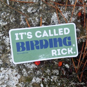 May include: A green sticker with white text that reads "It's called birding, Rick." The sticker is on a rock with brown twigs and red berries.
