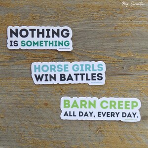 May include: Three white stickers with black and green text on a wooden surface. The stickers read: "NOTHING IS SOMETHING", "HORSE GIRLS WIN BATTLES", and "BARN CREEP ALL DAY, EVERY DAY."