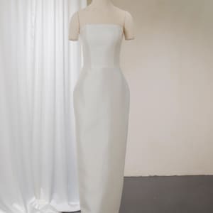 Könnte beinhalten: Ein trägerloses, bodenlanges weißes Kleid auf einer Schaufensterpuppe. Das Kleid hat ein eng anliegendes Oberteil und einen geraden Rock. Die Schaufensterpuppe steht vor einem weißen Vorhang und einer neutralfarbenen Wand.