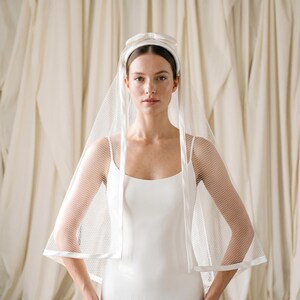 May include: A white bridal headband featuring a centered bow detail, paired with a shoulder-length veil made of fine white netting. The veil is finished with a smooth, solid white fabric trim along the edges, creating a clean and structured look.