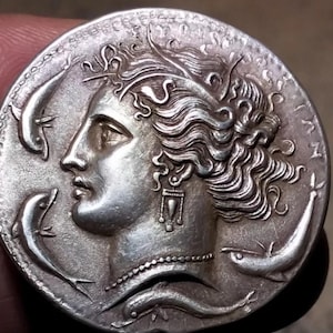 Puede incluir: Moneda de plata con un perfil de una mujer con el pelo largo y suelto y un collar. La moneda tiene tres diseños de peces alrededor del borde.