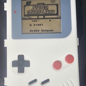 May include: A white Game Boy handheld video game console with a gray screen displaying the title "SUPER MARIOLAND" and the text "TOP- START 01989 Nintendo". The console has a black directional pad, two red buttons, and a speaker.