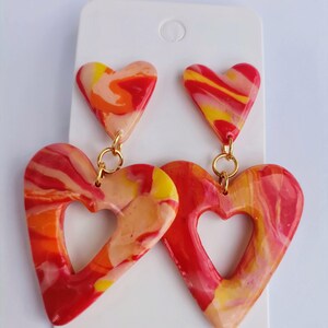 Puede incluir: Pendientes colgantes con dos piezas en forma de corazón cada uno. La pieza superior es un corazón sólido y la inferior es un marco de corazón abierto más grande. Los pendientes presentan un patrón jaspeado en tonos rojo, naranja, amarillo y blanco, unidos por anillas de metal dorado.
