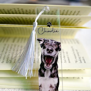 Könnte beinhalten: Transparenter Acryl-Lesezeichen mit einem lächelnden Hund, dem Namen "Charlie" und einem Pfotenabdruck. Das Lesezeichen hat eine weiße Quaste und befindet sich in einem offenen Buch.