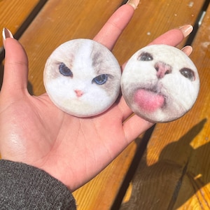 Könnte beinhalten: Zwei runde Katzen-Gesichts-Designs. Eines zeigt ein weißes und graues Katzen-Gesicht mit blauen Augen. Das andere hat ein weißes Katzen-Gesicht mit herausgestreckter Zunge. Die Katzen-Gesichter sind auf einem weichen, plüschigen Material gedruckt.