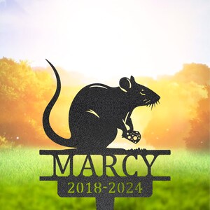Könnte beinhalten: Schwarzer Metall-Gartenstecker mit einer Ratten-Silhouette, die Käse hält. Der Stecker zeigt den Namen "MARCY" und die Jahre "2018-2024". Geeignet für den Außenbereich.
