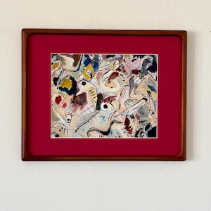 May include: Abstract watercolor painting in a wooden frame with a red mat. The artwork features a colorful composition of shapes and forms in shades of blue, yellow, brown, and white. The frame is a dark brown color.