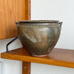 May include: A handcrafted, brown ceramic pot with a textured surface and two small handles. The pot has a wide opening and a slightly rounded shape. The color is a mix of brown and green, with a rustic, earthy appearance.