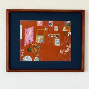 May include: A framed art print featuring a terracotta-coloured interior scene with various paintings on the walls, a table with a green bottle, a wine glass, and a chair. The artwork is enclosed in a dark wood frame with a deep navy blue matting border.