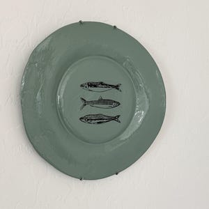 May include: A round, sage green ceramic plate with a glossy finish, featuring a centered black ink illustration of three stacked fish. The plate is secured to the wall with two small metal clips at the top and bottom edges of the rim.