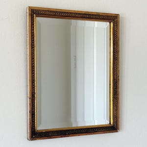 Puede incluir: Espejo rectangular con un marco dorado y marrón ornamentado. El marco presenta un patrón detallado a lo largo de los bordes. El espejo está colgado en una pared blanca, reflejando un vistazo de una habitación.