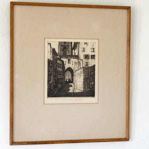 May include: A framed black and white etching depicting a narrow European street scene with stone buildings and a prominent arched stone gateway. The artwork is set within a light-colored mat and a simple, thin wooden frame with a natural finish.