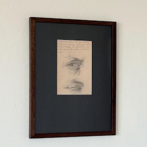 Puede incluir: Boceto a lápiz de un ojo y una boca, enmarcado en madera marrón oscuro. La obra de arte está sobre un fondo negro. Se ve texto manuscrito encima del ojo en el boceto. La obra enmarcada está montada en una pared blanca.