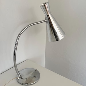 May include: A chrome desk lamp with a conical shade and a flexible gooseneck arm. The lamp has a round base and a series of small perforations on the shade. The lamp is on a white surface.