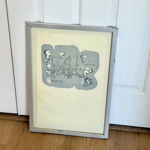 K&ouml;nnte beinhalten: Ein gerahmter Druck mit einer surrealen Illustration auf cremefarbenem Hintergrund. Eine graue Form enth&auml;lt schwarze Strichzeichnungen abstrakter Figuren und komplexer geometrischer Rohre. Der Text "...y paciente" und "Lunes de ocio" ist sichtbar.