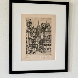 May include: A framed black and white etching depicting a historic street scene in Frankfurt, Germany, featuring tall timber-framed buildings and a cathedral spire in the background. The print includes handwritten text at the bottom.