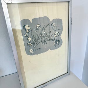 K&ouml;nnte beinhalten: Ein gerahmter Druck mit einer grauen abstrakten Form, schwarzen Strichzeichnungen von vier stilisierten Figuren und einer geometrischen Struktur. Der Text "Juegos de mesa" und "...y por dentro" ist sichtbar. Silberner Metallrahmen.
