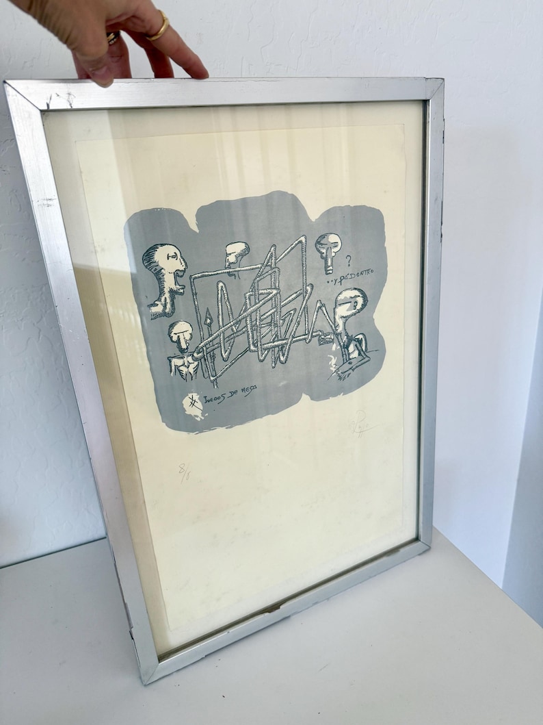 K&ouml;nnte beinhalten: Ein gerahmter Druck mit einer grauen abstrakten Form, schwarzen Strichzeichnungen von vier stilisierten Figuren und einer geometrischen Struktur. Der Text "Juegos de mesa" und "...y por dentro" ist sichtbar. Silberner Metallrahmen.