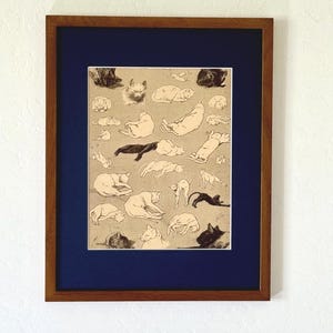 May include: A framed print featuring numerous cat illustrations in various poses. The artwork is set against a tan background and framed with a dark blue mat and a brown wooden frame. The cats are depicted in black and cream colors.