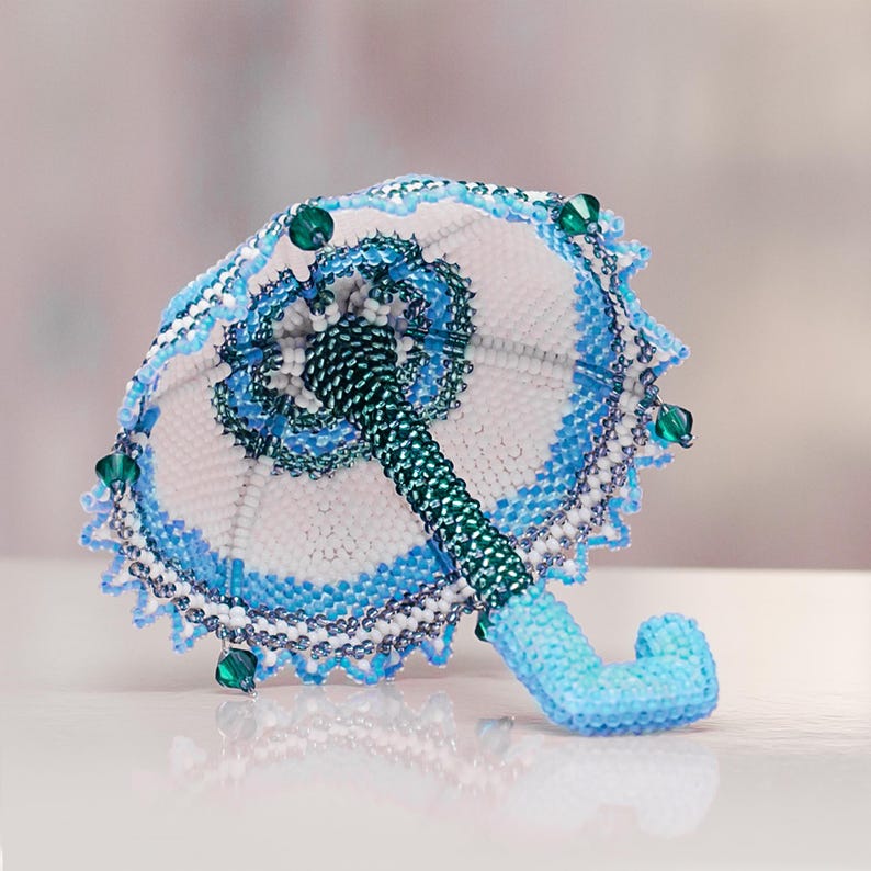 White and blue beaded umbrella in peyote stitch lies with the handle pointing forward. Tiny handmade beadwork accessory.