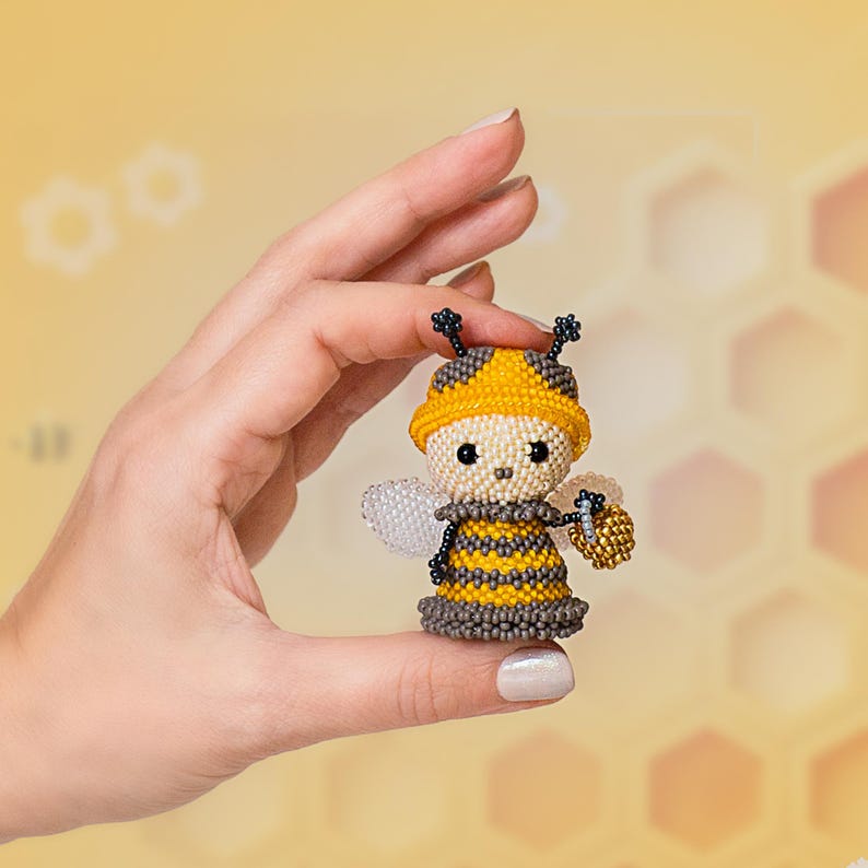 A girl gently holds a tiny beaded bee wearing a yellow hat and carrying a golden honey bucket, showcasing the miniature insect created with a Peyote Stitch pattern.