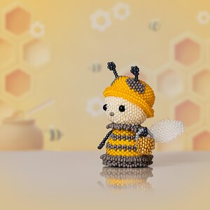 A Peyote Stitch beaded bee in a yellow hat stands turned to the left with a golden honey bucket, showing details of the beaded insect pattern.