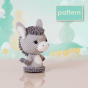 Könnte beinhalten: Eine kleine Eselfigur aus winzigen grauen und weißen Perlen. Der Esel hat einen runden Kopf, spitze Ohren und eine weiße Schnauze. Ein rechteckiges Schild in der oberen rechten Ecke zeigt das Wort pattern in weißer Schrift auf türkisfarbenem Grund.