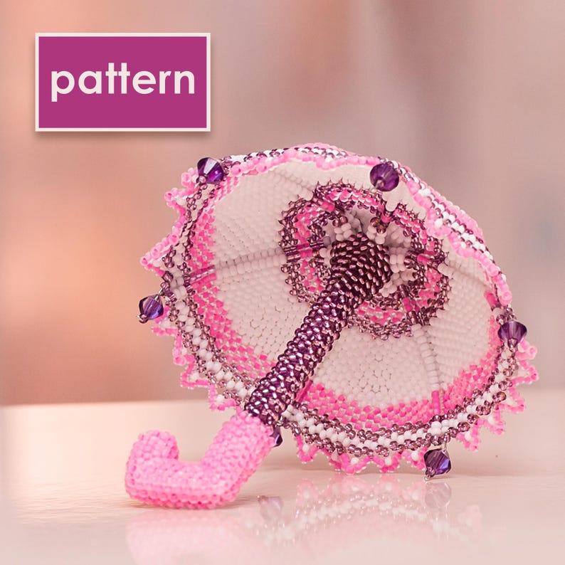 White and pink beaded umbrella made in peyote stitch lies with the handle pointing forward, showing the delicate beadwork pattern.