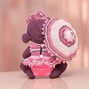 Back view of a beaded purple bear in a pink lace dress and bow headband sitting next to a white and pink umbrella. Peyote stitch beadwork toy.
