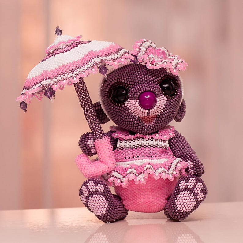 A lovely beaded bear figurine made with peyote stitch, wearing a pink dress and bow headband, holds a white and pink umbrella above her head. Front view.