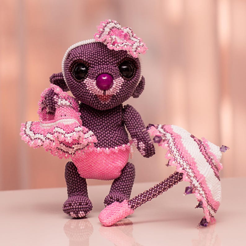 Purple beaded teddy bear in lace panties holding a tiny pink beaded dress in one hand, with a white and pink umbrella lying nearby. Peyote stitch handmade figurine.