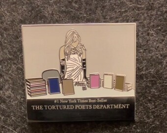 May include: A silver enamel pin depicting a woman standing behind a counter with a stack of books. The text on the pin reads "#1 New York Times Best-Seller THE TORTURED POETS DEPARTMENT".