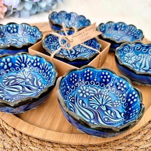 Może przedstawiać: Zestaw niebieskich ceramicznych misek z falistym brzegiem i skomplikowanymi wzorami kwiatowymi. Miski ułożone są na drewnianej tacy, niektóre w kartonowym pudełku. Miski mają ciemnoniebieską obwódkę i jaśniejsze niebieskie wnętrze, ok. 10 cm średnicy.