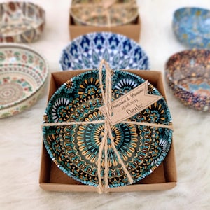 May include: A round ceramic bowl with a dark teal, orange, and black mandala pattern, packaged in a square brown cardboard box tied with twine. A small brown paper tag attached to the twine features the text Franziska & Dominic 15.08.2025 Danke.