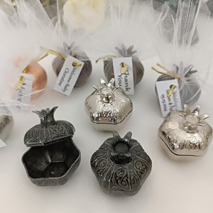 May include: An assortment of decorative pomegranate-shaped boxes in silver and dark gray. Some are open, revealing the interior. Several are adorned with tags and tulle. A bouquet of white and green flowers is in the background.