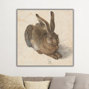 May include: A detailed pencil drawing of a brown hare lying on its side, with its head turned to the left. The hare's fur is depicted with great detail, and its long ears are visible. The drawing is on a light beige background.