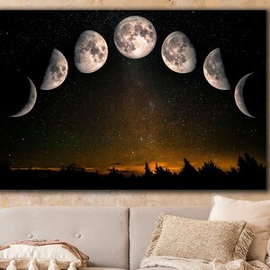May include: A canvas print of a crescent moon phases design against a starry night sky. The moon phases are in a semi-circle shape with the full moon in the center. The print is hanging on a wall above a light gray couch with pillows and a throw blanket.