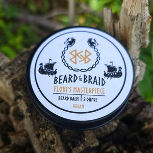 May include: A round, black and white tin of beard balm. The lid features the text "BEARD & BRAID" and "FLOKI'S MASTERPIECE". The tin also includes a Viking ship illustration and is labeled "2 OUNCE" and "VEGAN".
