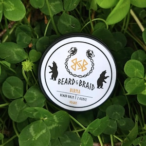 May include: A round black and white container of beard balm, with the text "BEARD & BRAID" and "BIRNA" printed on the label. The label also features bear and Viking-inspired graphics. The container is 2 ounces.