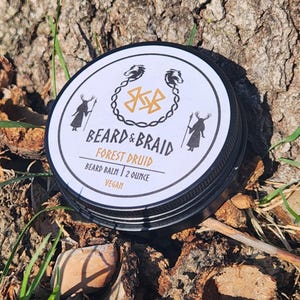 May include: A round tin of Beard & Braid Forest Druid beard balm. The label is white with black text and illustrations. The tin is black with the text "2 ounce" and "Vegan". The tin is sitting on a bed of wood and leaves.