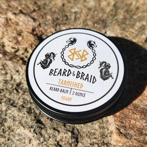 May include: A round, black and silver tin of beard balm. The white lid features the words "BEARD & BRAID" and "TARNISHED" in a stylized font. The label also includes the text "BEARD BALM | 2 OUNCE" and "VEGAN".