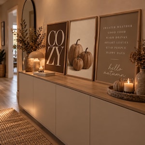 K&ouml;nnte beinhalten: Drei gerahmte Wandbilder auf einem Sideboard. Das linke Bild zeigt das Wort COZY in gro&szlig;en wei&szlig;en Buchstaben auf dunklem Grund. Das mittlere Bild zeigt drei K&uuml;rbisse. Das rechte Bild listet herbstliche Begriffe und den Text hello autumn auf.